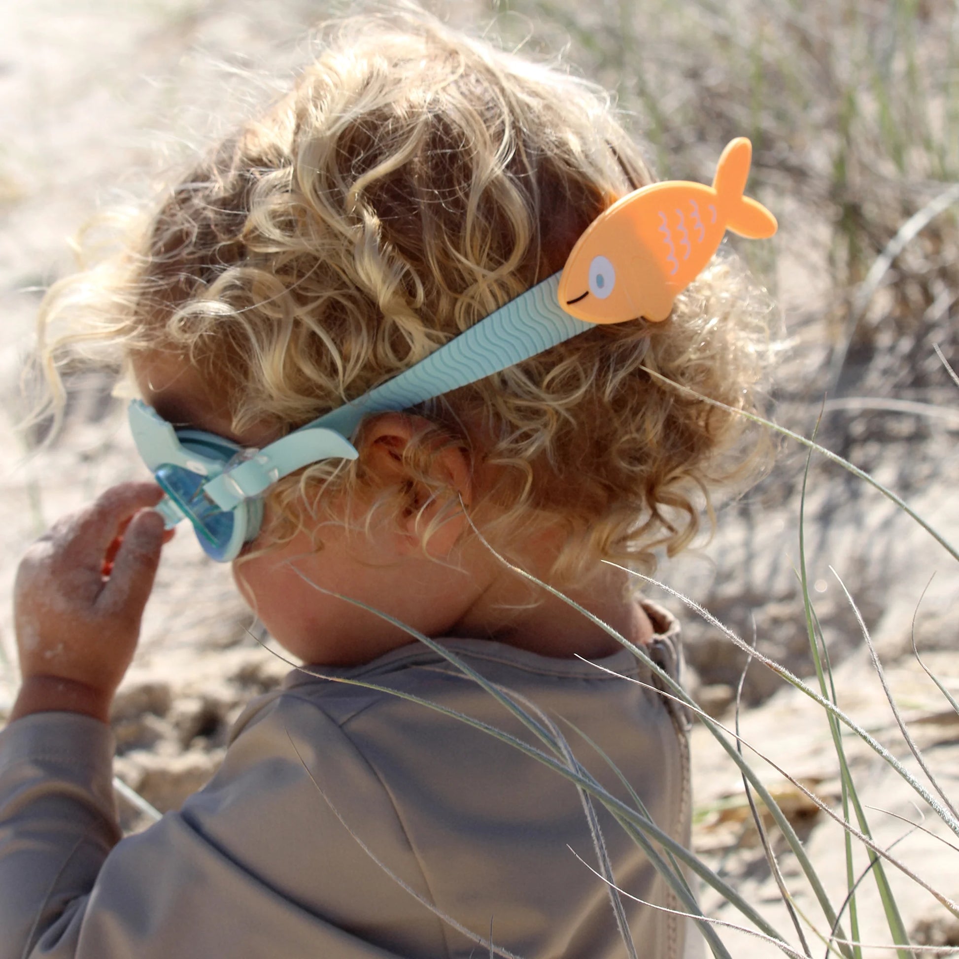 A child with curly hair sits on a sandy beach, wearing a gray shirt and Sunnylife's Finn the Fish Aqua Blue Swimming Goggles, ready for underwater play.