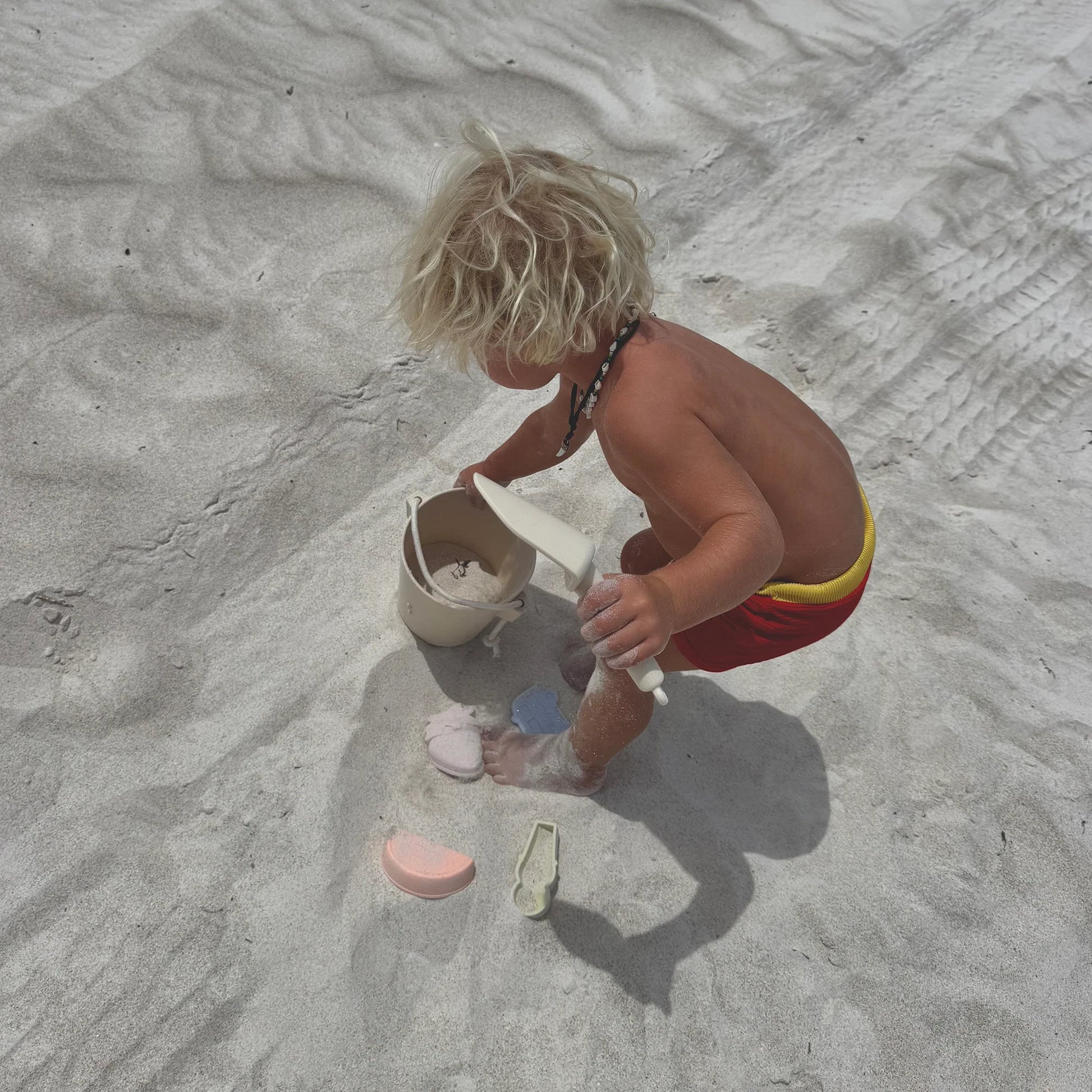A young child with blond hair and red shorts plays on light-colored sand, enjoying water play with the Sunnylife Silicone Bucket & Spade Set Apple Sorbet Multi and beach toys.