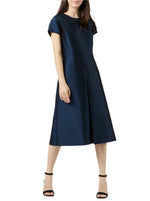 A woman wears the Ann Mashburn Suzzette Dress, a navy blue knee-length pleated A-line with cap sleeves, paired with black high-heeled sandals, standing against a white background.