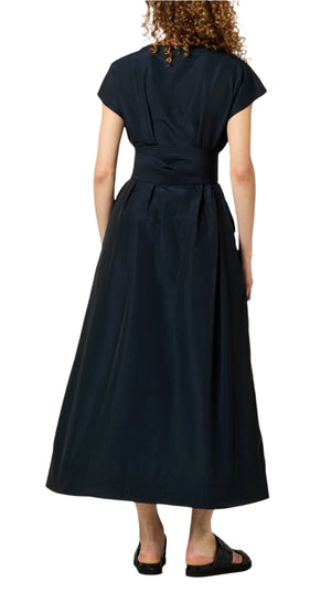 The person is wearing the Gigi Dress by Ann Mashburn, an elegant black ankle-length dress with short sleeves and a wrap belt, featuring subtle trapunto stitching, viewed from behind.