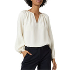 A woman models the Ariana Blouse by Ann Mashburn, showing its long sleeves, off-white crepe de chine fabric, and v-neckline, paired with black trousers and posing with one hand in her pocket against a plain background.