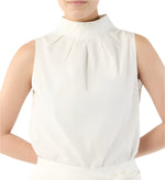 A person wearing the Ann Mashburn Clair Top, a white sleeveless mock neck tank with a high pleated neckline, stands with hands behind their back.