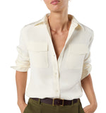 A person wearing the Hannah Blouse by Ann Mashburn, an off-white utilitarian top with chest pockets, paired with olive green pants and hands in pockets.