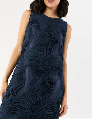 A woman stands against a plain background wearing the Ann Mashburn Susy Shift Maxi Dress, a sleeveless navy blue dress featuring textured, wavy fabric patterns.