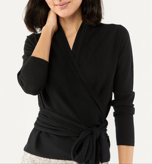A woman wearing the Ann Mashburn Mika Cashmere Wrap Sweater, a luxury black long sleeve top with a tied waistband, stands with one hand touching her neck.