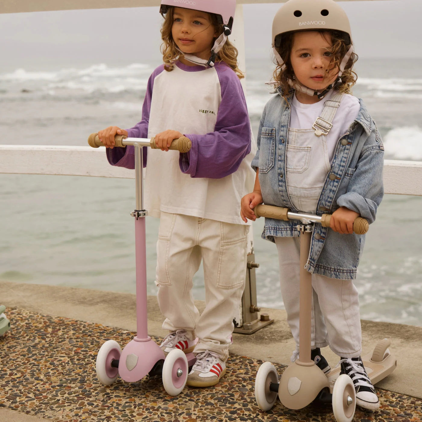 Two young children wearing helmets stand outdoors by a white fence and the ocean, riding Banwood Eco Scooters made from recycled plastic for eco-friendly fun. They’re dressed in casual clothes and sneakers.