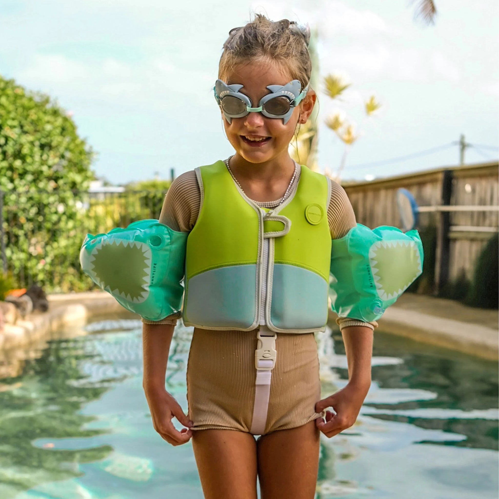 A confident child in their green and blue life jacket and Sunnylife Salty the Shark aqua-themed goggles stands proudly by the pool, ready to splash around with their trusty Sunnylife Float Bands Salty the Shark Aqua.