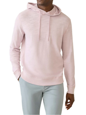A person in a relaxing Faherty Sunwashed Slub Hoodie in light pink and relaxed-fit light gray pants stands casually with one hand in their pocket.