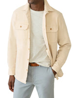 A man wears the Faherty Sunwashed Canvas CPO, a relaxed fit light beige shirt jacket, over a white and gray striped tee with light gray pants and a brown belt.