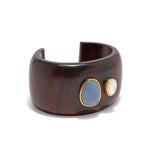 Introducing the Lanna Cuff by Lizzie Fortunato: a stunning acacia wood bracelet accented with two gemstones—a large blue and a smaller white—set in gold-plated brass for timeless elegance.
