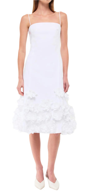 A woman in a Simkhai Bella Floral Dress with floral hem accents, paired with white shoes, stands against a plain background.