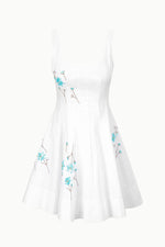 The Staud Mini Wells Dandelion Dress is a white sleeveless dress featuring a flared skirt, blue floral embroidery on the bodice and sides, and a subtle corset-style panel for a flattering fit.