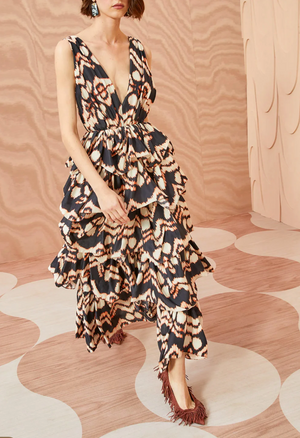 A woman wears the Ulla Johnson Dephne Dress, a sleeveless silk taffeta midi with bold black, white, and tan geometric patterns and a ruffled skirt, standing on a decorative floor with matching wavy wall panels.