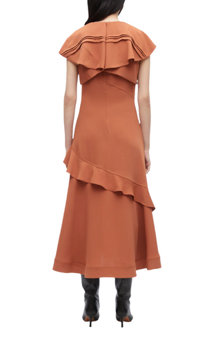 A woman, exuding romance, faces away in a rust-colored Ruffle Overlay Midi Dress from 3.1 Phillip Lim with an asymmetrical hem and layered ruffles. She pairs it with black heeled boots for an edgy touch to her elegant ensemble.
