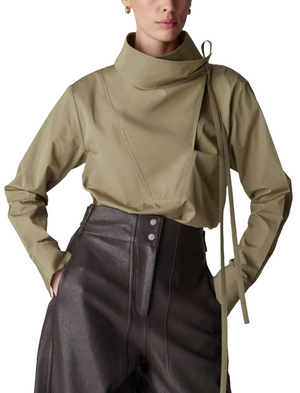 A person is wearing CO's Fencer's Fluid Blouse, a stylish beige high-neck top with tie detail, paired with high-waisted dark pants featuring a subtle flyaway hem for added flair.