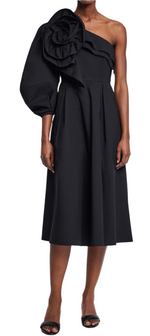 Wearing the Dorothee Schumacher Poplin Power Flower Dress, a black one-shoulder midi with a large rosette and puffed sleeve, the individual elegantly pairs it with black heels.