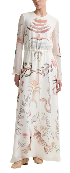 A person elegantly wears the Altuzarra Sybbeline Dress, a long sheer silk piece adorned with colorful abstract designs and text, paired with beige shoes. The dress's maxi hem gracefully touches the floor against a plain background.
