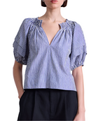 A woman wears the Apiece Apart Caminitas Puff Sleeve Top—a blue and white vertical striped blouse with puffed sleeves and a V-neck—paired with black pants, posing against a white background.