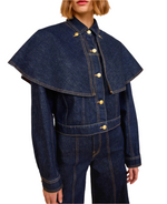 A person wearing the Ulla Johnson Agata Denim Cape Jacket, a vintage-inspired dark denim piece with a wide cape-like collar, paired with matching denim pants, shown from mid-torso up.