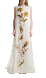 A woman wears the Altuzarra Jools Dress, a sleeveless, floor-length white silk gown featuring a vertical botanical print with large yellow and purple thistle flowers and green stems.
