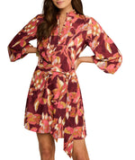 A woman wears the Figue Izzie Dress, a maroon matte crepe piece with pink, orange, and white abstract floral prints, long sleeves, a belted waist, and an A-line knee-length silhouette.
