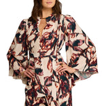 A woman wears the Avery Top by Figue—a long-sleeve, patterned viscose crepe blouse featuring a deep V-neck, tie waist, and butterfly sleeves in red, black, and cream.