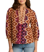 A woman wears the Figue Danilia Top—a relaxed fit shirt with deep V-neck, ruffled collar, and maroon, orange, and white geometric print—paired with light blue jeans.