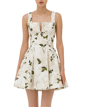 A woman models the Agua Dress by Agua By Aguabendita, a sleeveless white mini dress with green leaf patterns and a fitted bodice, featuring an elegant sweetheart neckline.