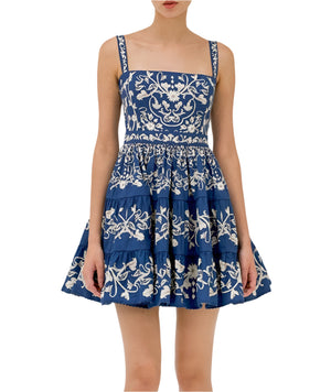 A woman wears the Lima Mini Dress by Agua By Aguabendita, a blue sleeveless cotton mini dress with white floral embroidery and a tiered skirt, standing against a plain background.