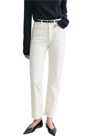 A person wears Toteme’s Classic Cut Denim in white organic cotton, styled with a black sweater, black belt, and pointed black shoes, posing with one hand in their pocket against a white background.