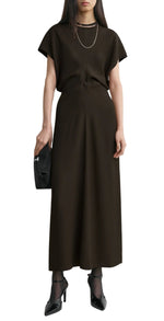 A woman wears Toteme's Slouch Waist Dress in black with short sleeves and a floor-length hem, styled with black heels, sheer socks, a necklace, and a black clutch.