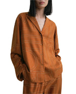 A woman wearing a burnt orange Toteme Monogram Silk PJ Top with a geometric line pattern and matching silk twill pants, standing with hands in her pockets.