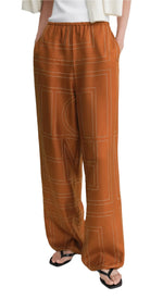Someone wearing Toteme's Monogram Silk PJ Bottoms—rust-colored silk twill pants with a geometric stitched pattern—paired with black sandals and a white top draped over their shoulders.