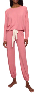 A woman wearing the Eberjey EBJ Gisele Slouchy Set—loose-fitting, long-sleeve pink pajamas made from sustainable TENCEL™ Modal, featuring a white waist tie and gathered ankle cuffs—stands barefoot in ultimate comfort.