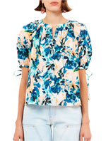 A person in Ulla Johnson's Vienne Blouse, a short-sleeve floral print in blue and pink hues made from 100% cotton, paired with light blue pants.