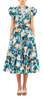 Someone wearing the Ulla Johnson Ottilie Dress, which features a floral print with blue, green, and peach hues, short sleeves, and a belted waist, paired with strappy sandals.