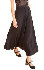 A person wearing the Ulla Johnson Rowan Skirt, a black A-line, fixed waist midi skirt with front buttons, paired with black heeled Mary Jane shoes and a sheer black top featuring floral embellishments.