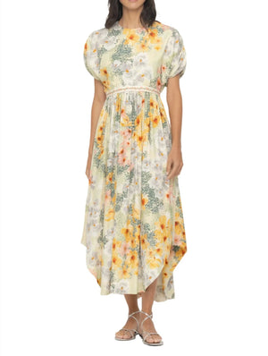 A woman wearing the Aire Maxi Dress by Agua By Aguabendita, a light floral A-line dress with puff sleeves and an asymmetrical hem, paired with silver sandals.