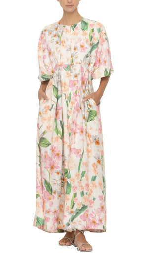 Wearing the Gesto Maxi Kaftan from Agua By Aguabendita, adorned with pink and green floral patterns, paired with white sandals, and hands in pockets exudes effortless charm.