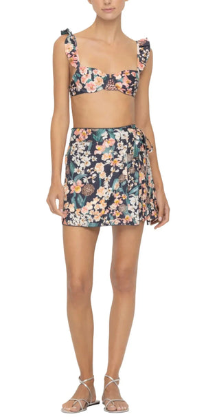A person wearing a floral bikini top with shoulder ruffles and the Luna Mini Pareo, elegantly tied at the waist, exudes a carefree vibe perfect for sunny days by the water. This Agua By Aguabendita ensemble is ideal for a fun day out.