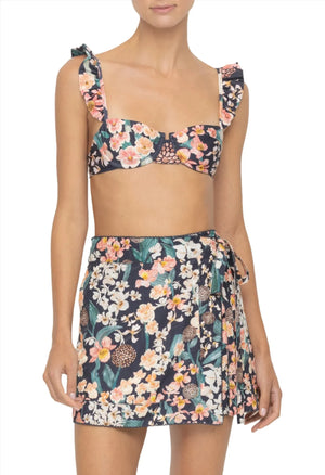 A person is wearing a floral bikini top with ruffled straps and a matching wrap skirt, complemented by the chic Luna Mini Pareo by Agua By Aguabendita, tied at the waist for an effortlessly stylish look.