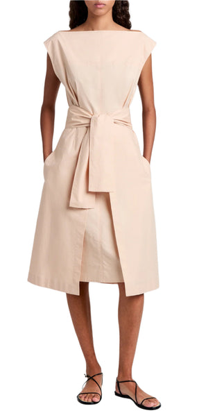 A person is dressed in a beige, sleeveless Proenza Schouler White Label Caro Dress in brushed poplin, featuring an A-line silhouette and self-tie waist belt, paired with black sandals.