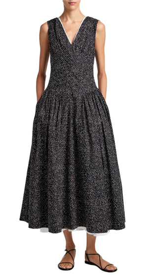 A person in a sleeveless Clayton Dress Printed Poplin by Proenza Schouler White Label, featuring a white dotted pattern, stands against a light background wearing black sandals with hands in pockets.