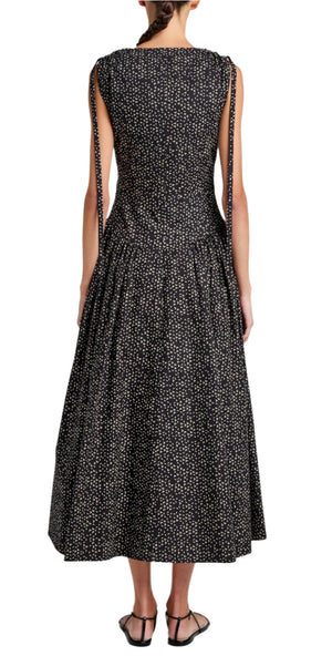 A person stands with their back to the camera wearing a black Proenza Schouler White Label Clayton Dress Printed Poplin with white speckles, sleeveless and tied shoulders. The ankle-length dress pairs elegantly with flat black sandals.