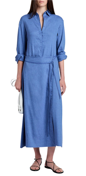 A person is wearing the Proenza Schouler White Label Lato Wrap Dress Linen Slub, a long blue button-up style with a shirtdress silhouette and belt, holding a white clutch. The sleeves are rolled up, and they complement it with black sandals for a chic finish.