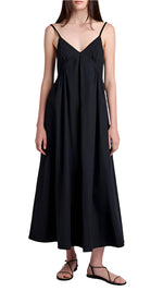 A woman in the Proenza Schouler White Label Barker Dress Stretch Poplin—a sleeveless black maxi with slim straps—pairs it with black flat sandals, standing against a white background.