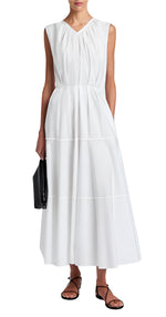 A woman wears the Kailey Dress Stretch Poplin by Proenza Schouler White Label—a sleeveless white V-neck pleated dress—paired with black strappy sandals and a black clutch.