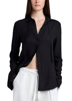 A woman models the Proenza Schouler White Label Cami Shirt Crinkle Viscose in black with long sleeves and white pants, hands in pockets, against a white background, highlighting its relaxed silhouette.