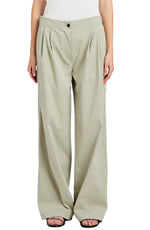 A person wears the Proenza Schouler White Label Romina Pant—loose-fitting, high-waisted, light beige cotton pants with a wide-leg silhouette, front pleats, black button closure—paired with open-toe black sandals.