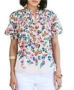 A woman models the Figue Wilma Top, a short-sleeved floral blouse with a split neckline and pintucked yoke in lightweight cotton voile, styled with white pants and gold bracelets.